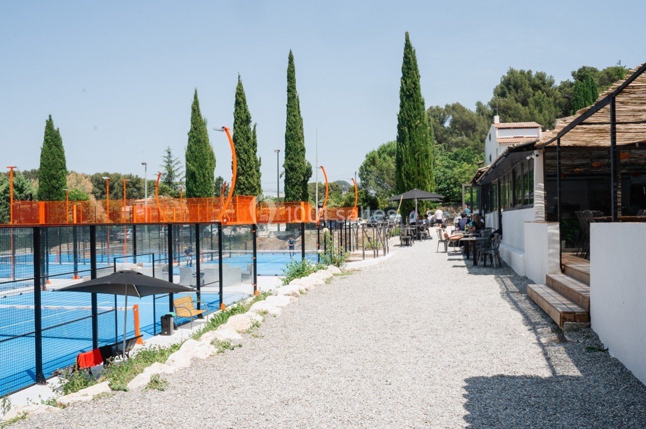 Terrasse en gravier avec tables et parasols, bordant des terrains de padel entourés de grillages et de cyprès.