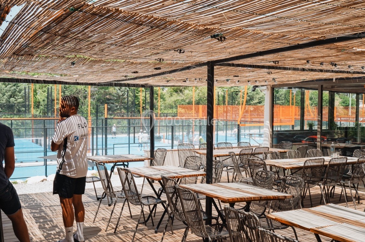 Terrasse ombragée avec tables en bois et chaises en métal, vue sur une piscine entourée de végétation.