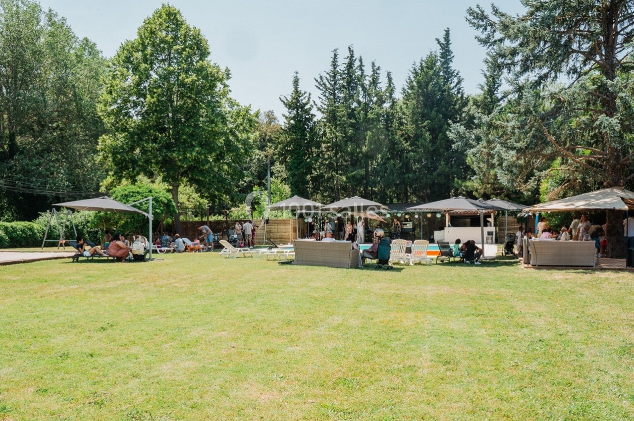 Espace extérieur avec pelouse, parasols, salons de jardin et personnes profitant d'un cadre arboré par temps ensoleillé.