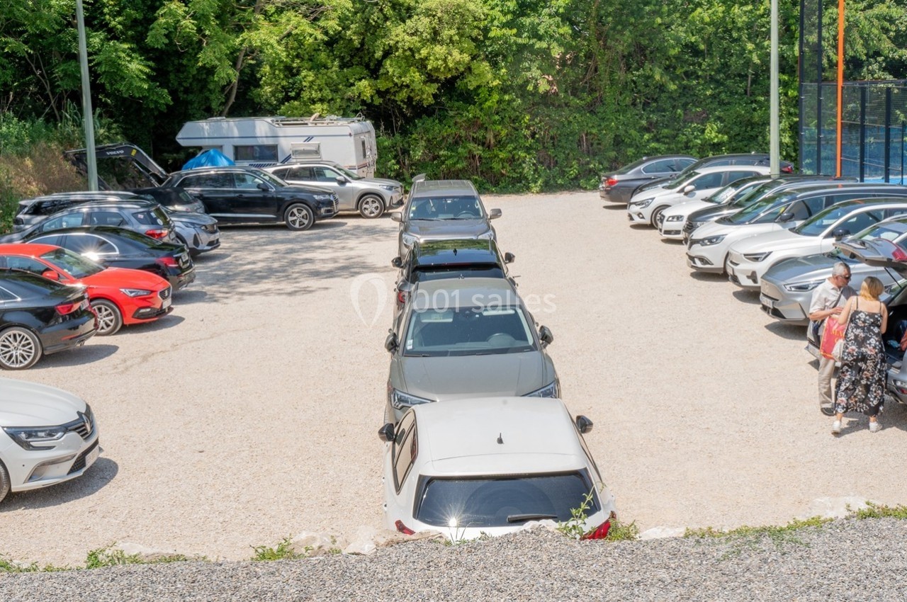Parking extérieur avec plusieurs voitures stationnées, entouré de végétation et d'un terrain de sport clôturé.