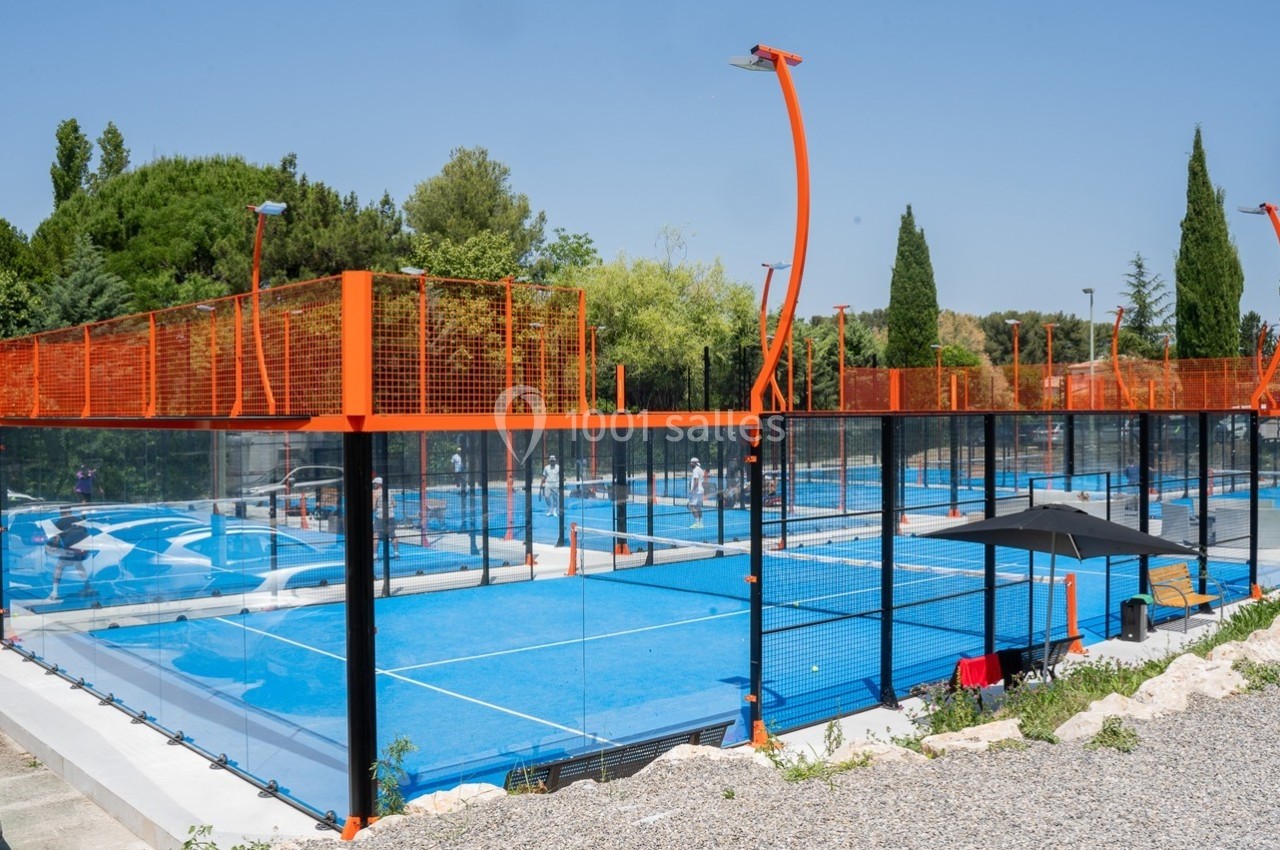 Terrains de padel extérieurs avec revêtement bleu, entourés de grillages orange, situés dans un espace arboré.