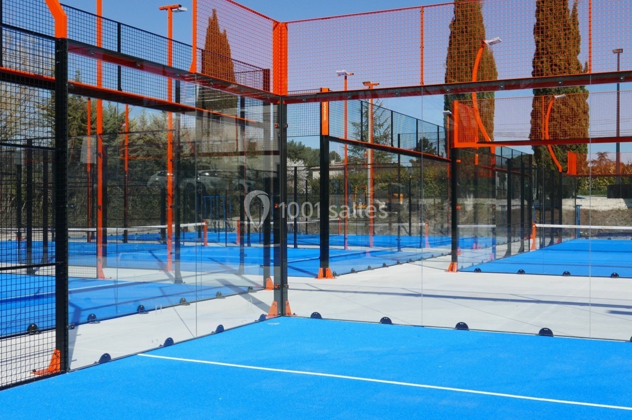 Terrains de padel extérieurs avec sol bleu, entourés de grillages et de parois vitrées, sous un ciel dégagé.