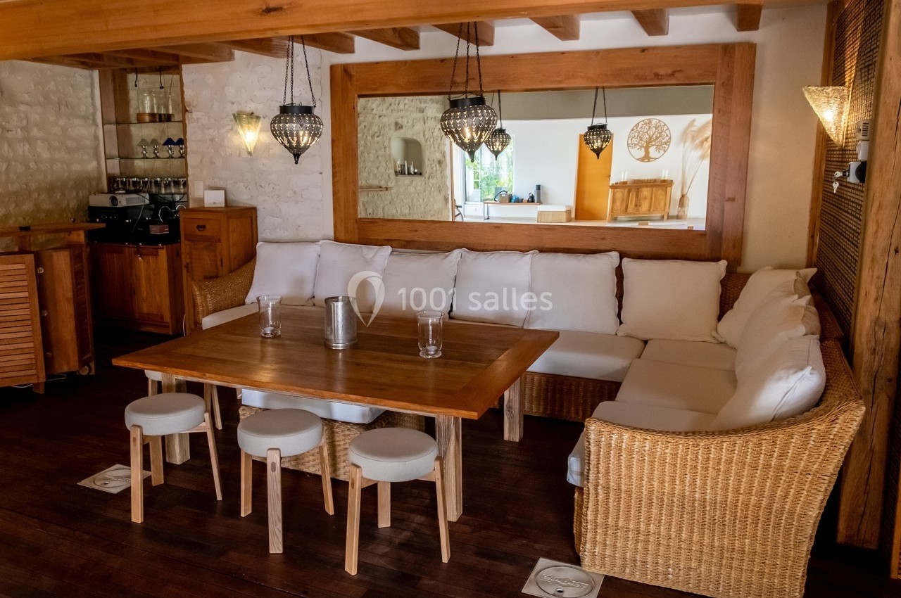 Salon chaleureux avec canapé en rotin, table en bois, tabourets et éclairage suspendu dans un intérieur rustique.