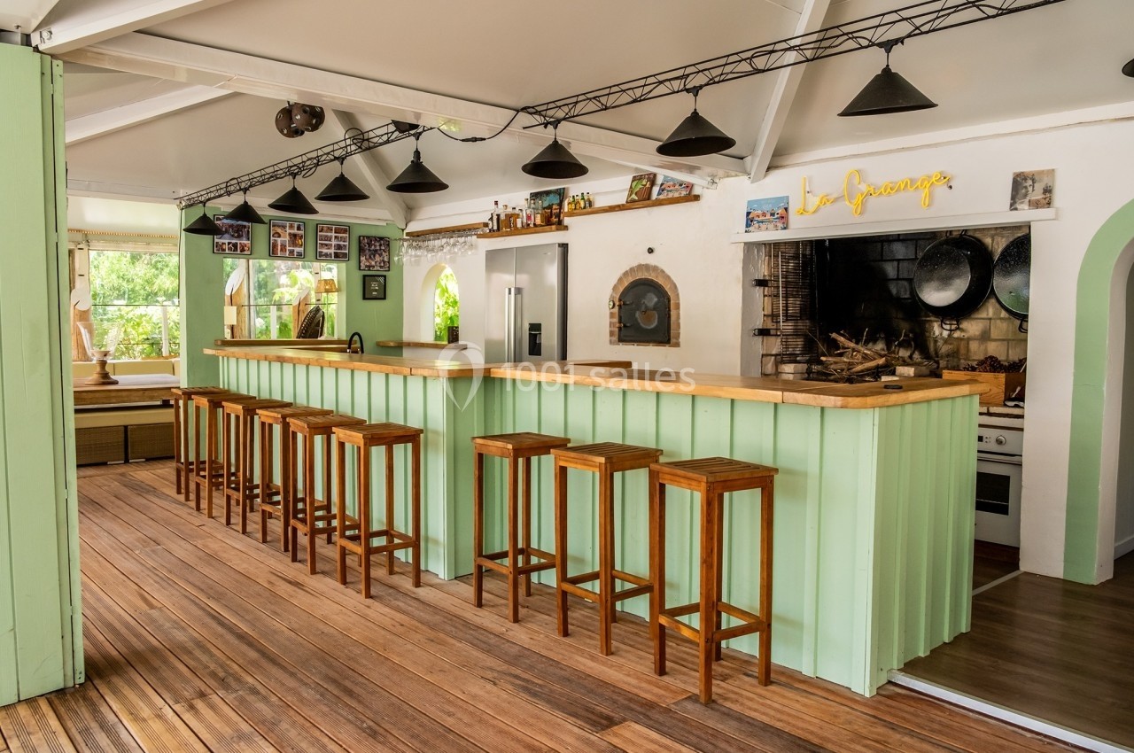 Cuisine ouverte avec un long comptoir en bois, tabourets alignés, éclairage suspendu et four à bois en arrière-plan.