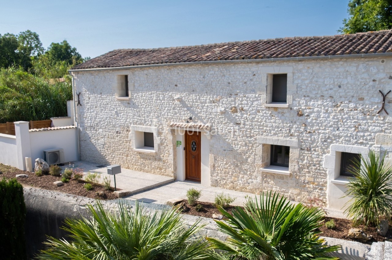 Façade d'une maison en pierre blanche avec toit en tuiles, entourée de végétation et d'un petit jardin.
