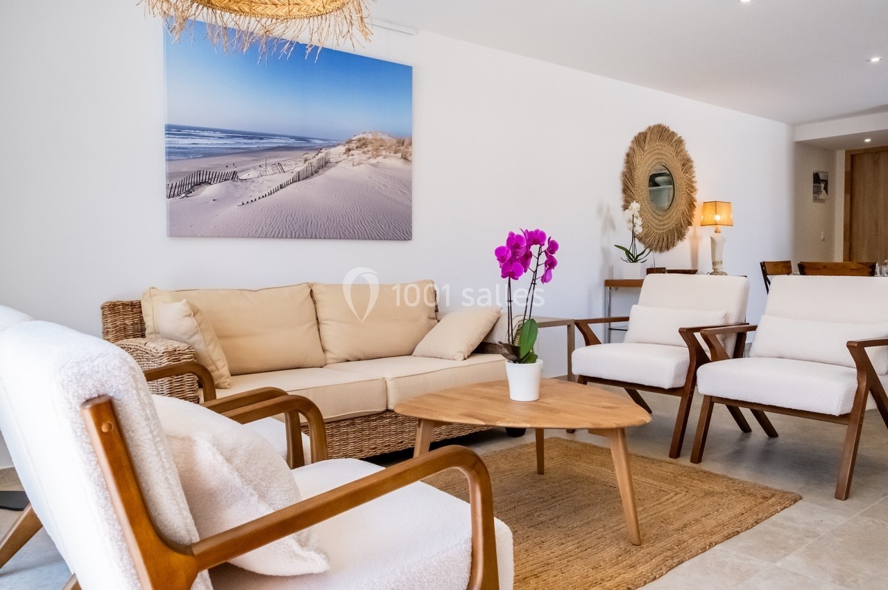 Salon lumineux avec mobilier en bois et rotin, orchidée violette sur une table basse, et tableau de dunes au mur.