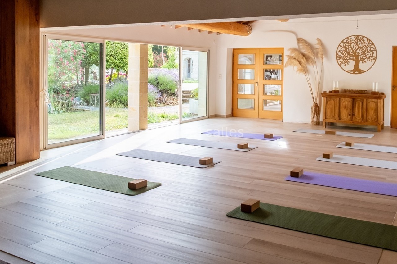 Salle lumineuse aménagée pour le yoga, avec tapis alignés, briques en bois et vue sur un jardin fleuri.