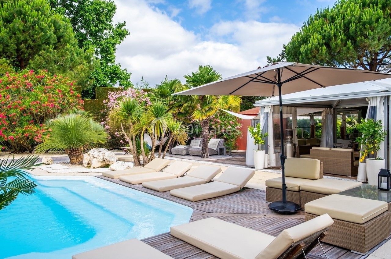Terrasse avec piscine, transats beige, parasol et végétation luxuriante sous un ciel partiellement nuageux.