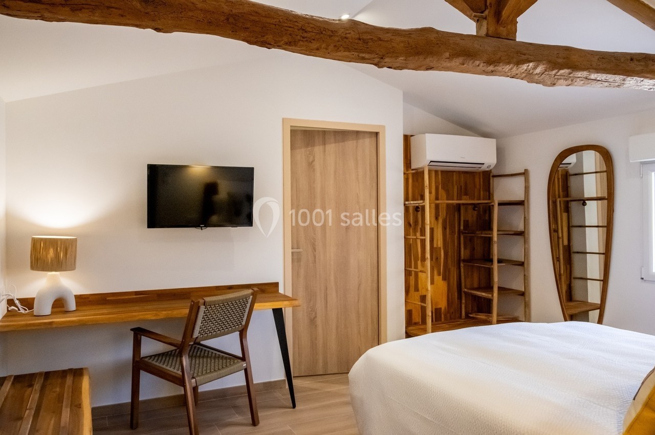 Chambre lumineuse avec lit, bureau en bois, chaise, télévision murale, étagères ouvertes et poutres apparentes.