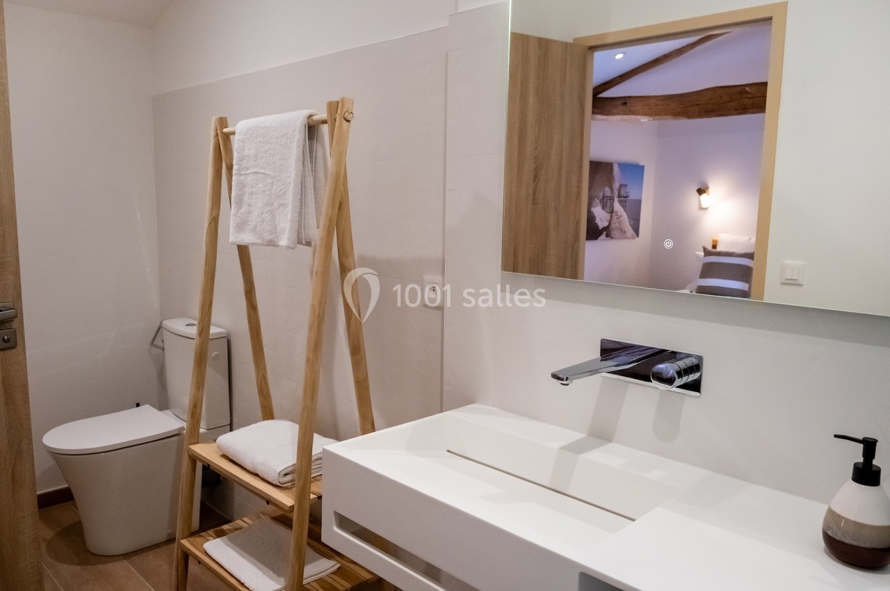 Salle de bain moderne avec lavabo blanc, miroir, porte-serviettes en bois et toilettes, éclairée par une lumière douce.