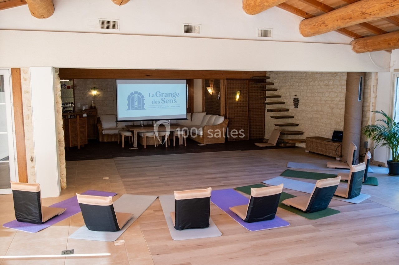 Salle lumineuse avec tapis de yoga, chaises de méditation et écran affichant ’La Grange des Sens’.
