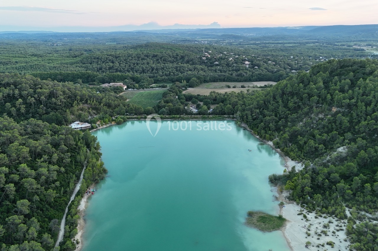 Vue aérienne d'un lac entouré de forêts et de collines verdoyantes sous un ciel légèrement nuageux.