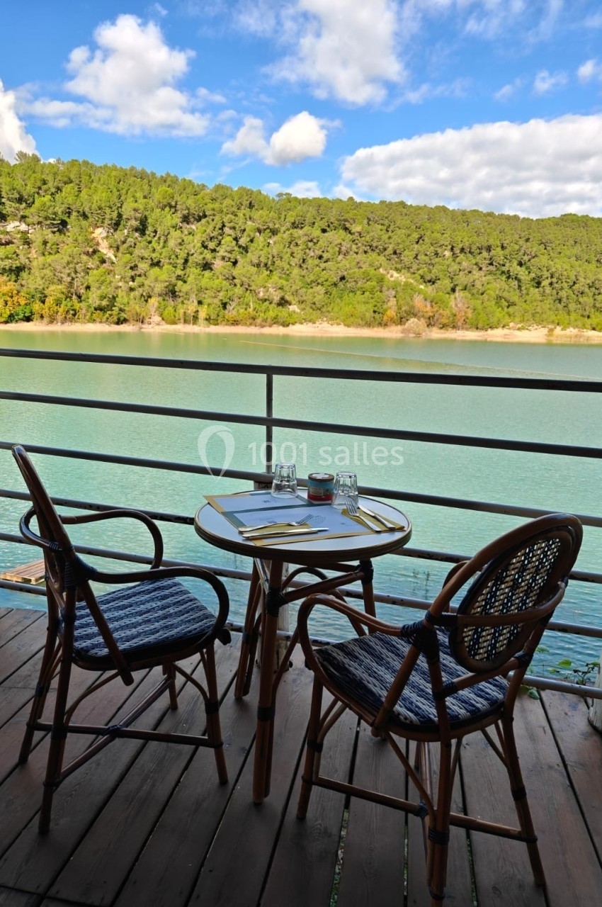 Table et deux chaises en rotin sur une terrasse en bois, avec vue sur un lac entouré de collines boisées.