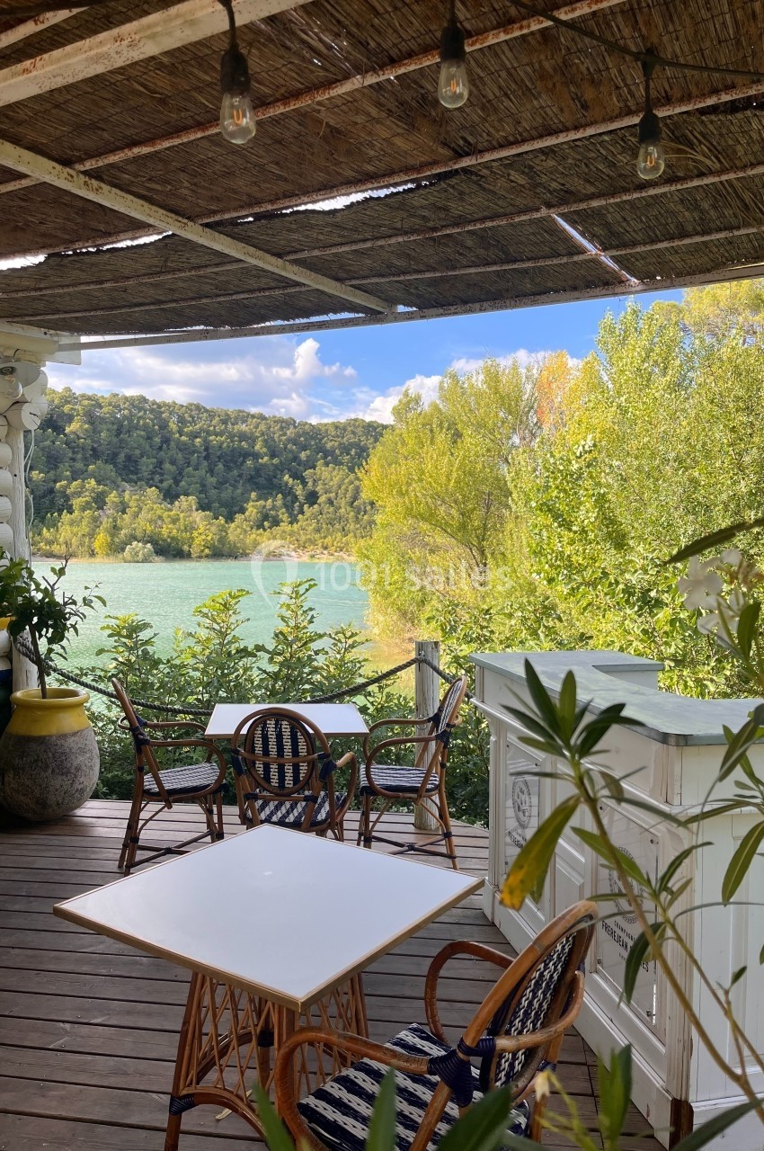 Terrasse en bois ombragée avec tables et chaises, vue sur un lac entouré de végétation et collines verdoyantes.