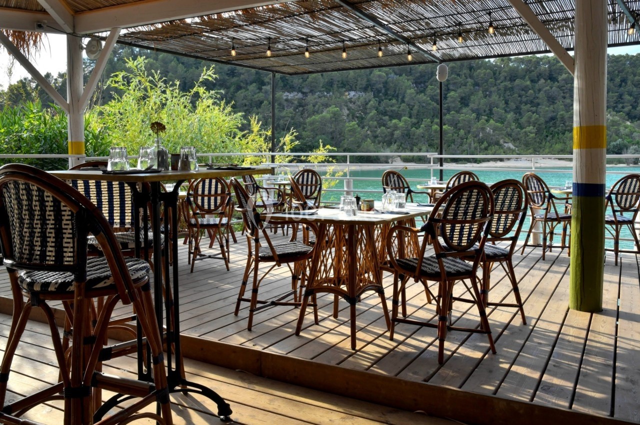 Terrasse en bois avec tables et chaises en rotin, surplombant un lac entouré de végétation et de collines.