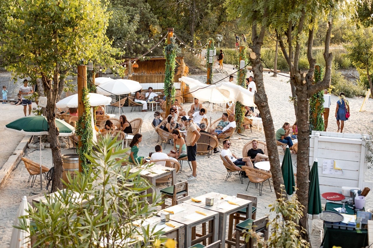 Terrasse en plein air avec des tables, des chaises en rotin et des parasols, entourée d'arbres et de végétation.