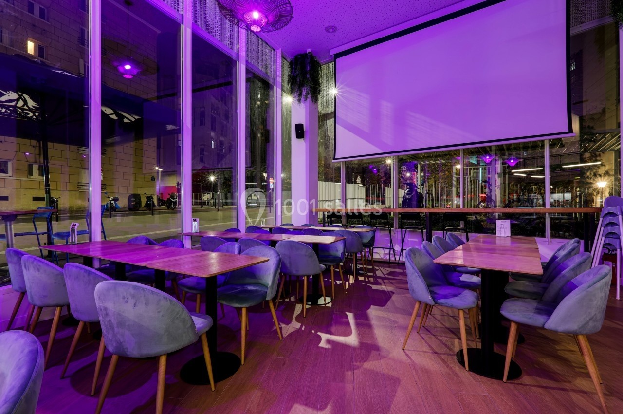 Salle de restaurant moderne avec chaises en velours, tables en bois et éclairage violet, vue sur l'extérieur vitré.