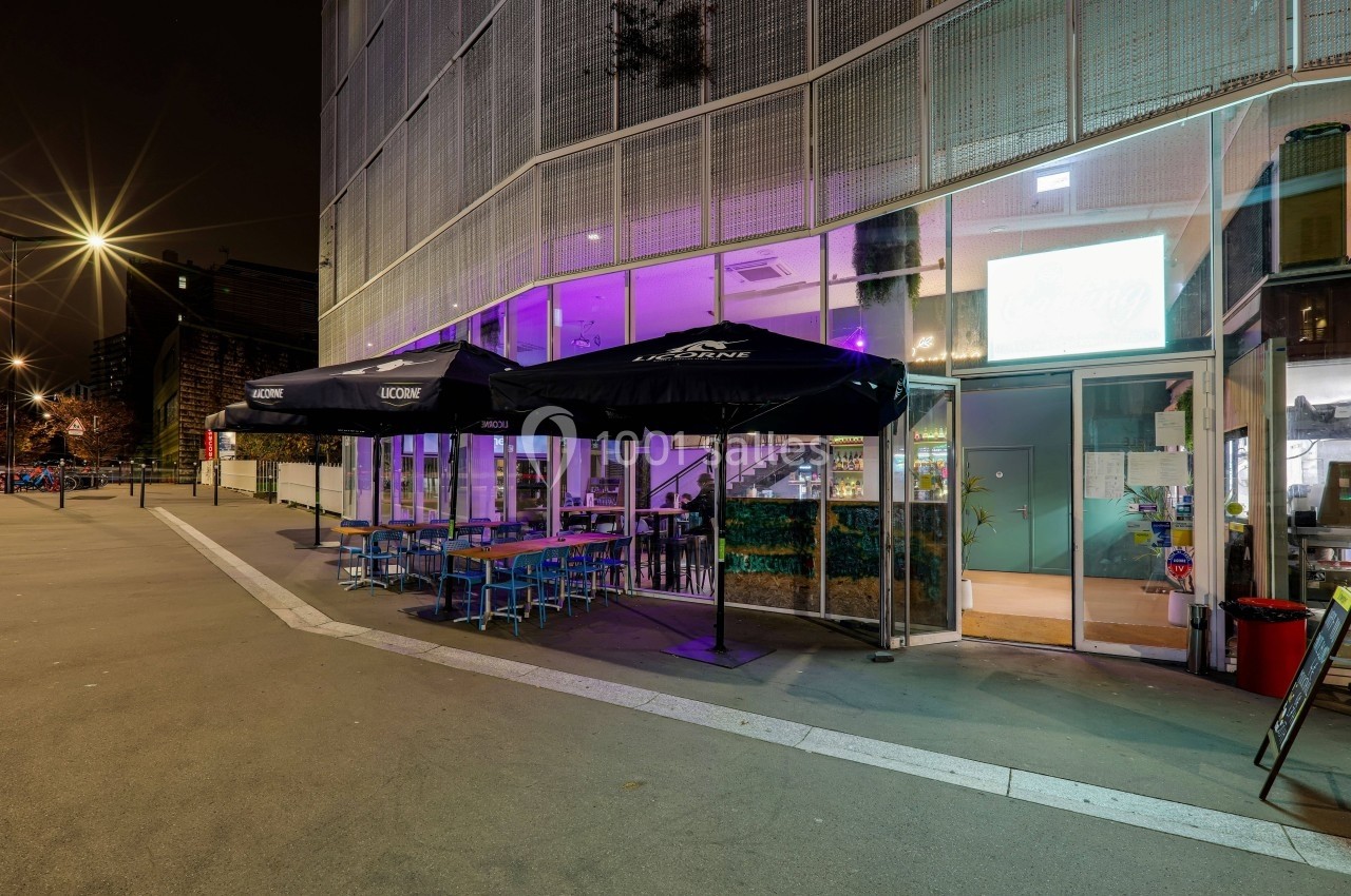 Terrasse d'un café-restaurant avec tables et chaises sous des parasols noirs, éclairée par des lumières nocturnes.