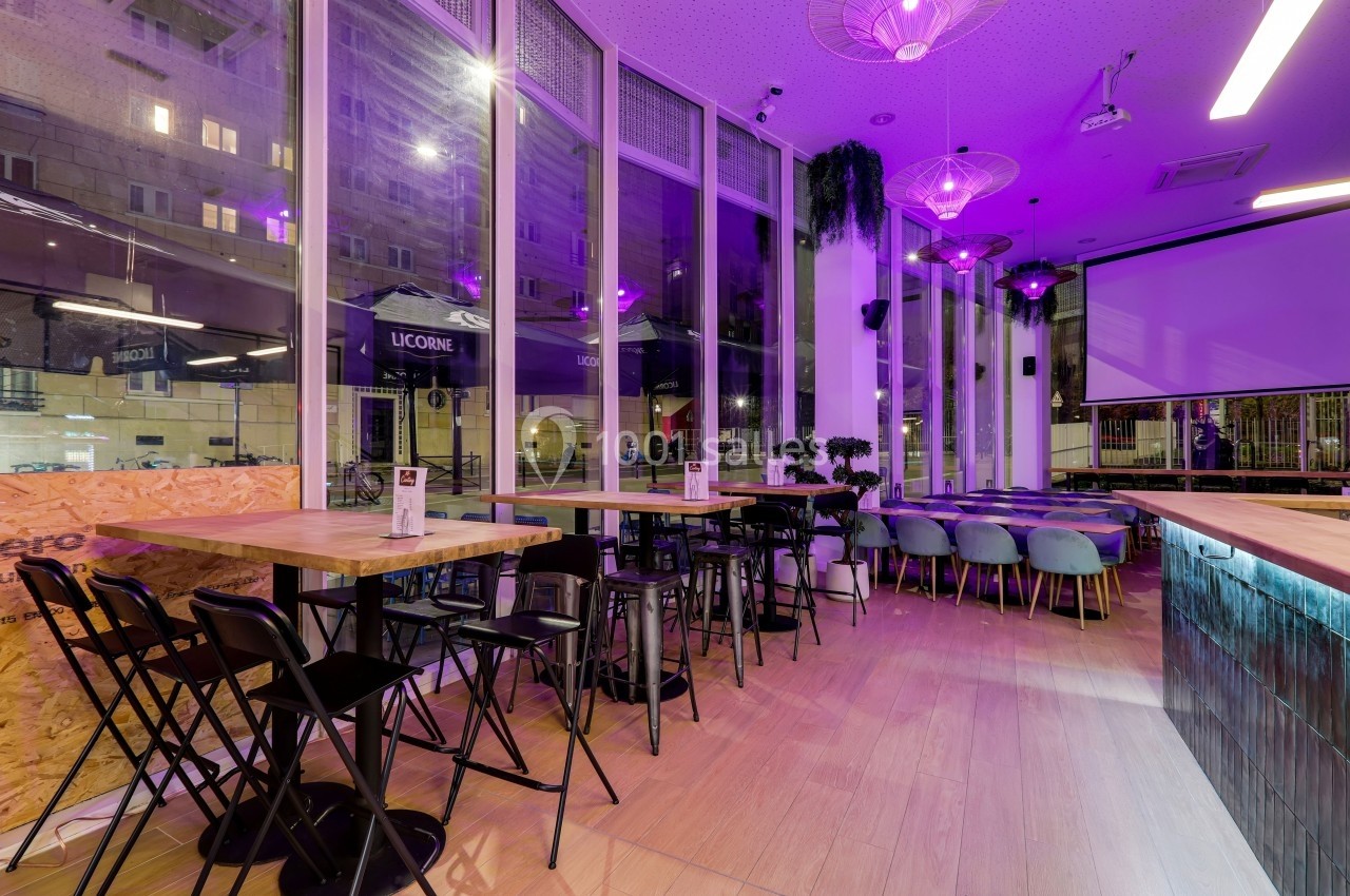 Intérieur d'un café moderne avec tables en bois, chaises noires et bleues, éclairage violet et grandes baies vitrées.