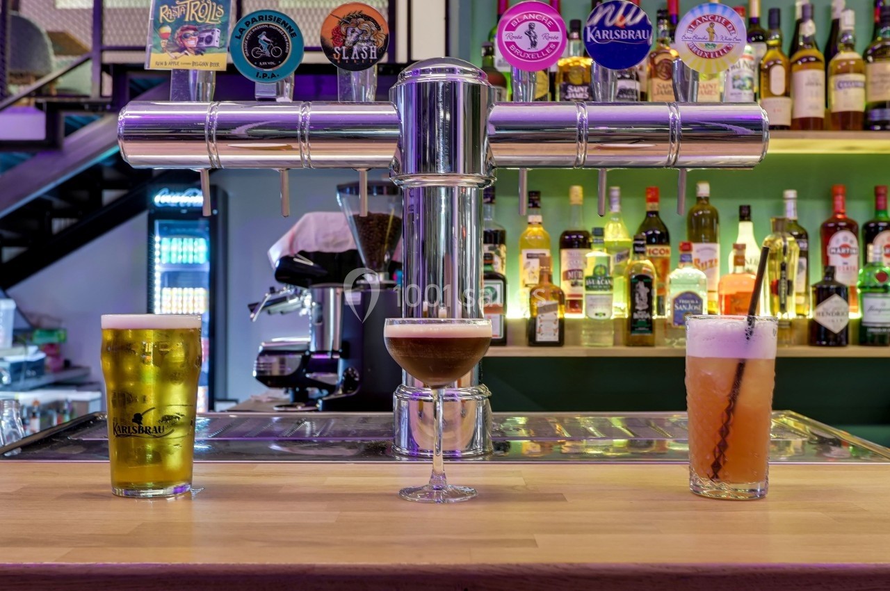 Trois verres de boissons variées posés sur un comptoir de bar avec des bouteilles et des tireuses en arrière-plan.