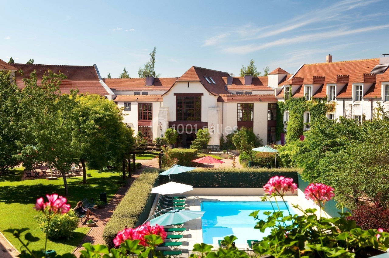 Vue d'un hôtel avec piscine extérieure, jardin verdoyant et bâtiments aux toits de tuiles rouges par temps ensoleillé.