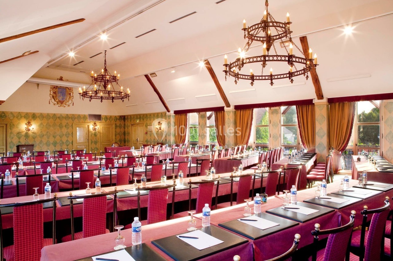 Salle de conférence lumineuse avec tables alignées, chaises rouges, bouteilles d'eau et carnets, décorée de lustres et…