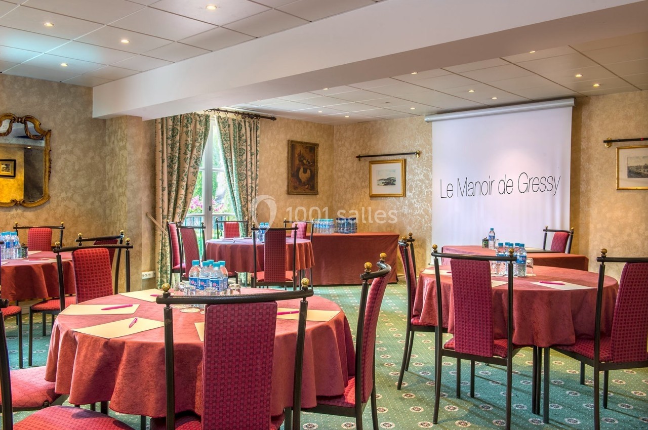 Salle de réunion avec tables rondes dressées, chaises rouges, écran de projection et lumière naturelle.