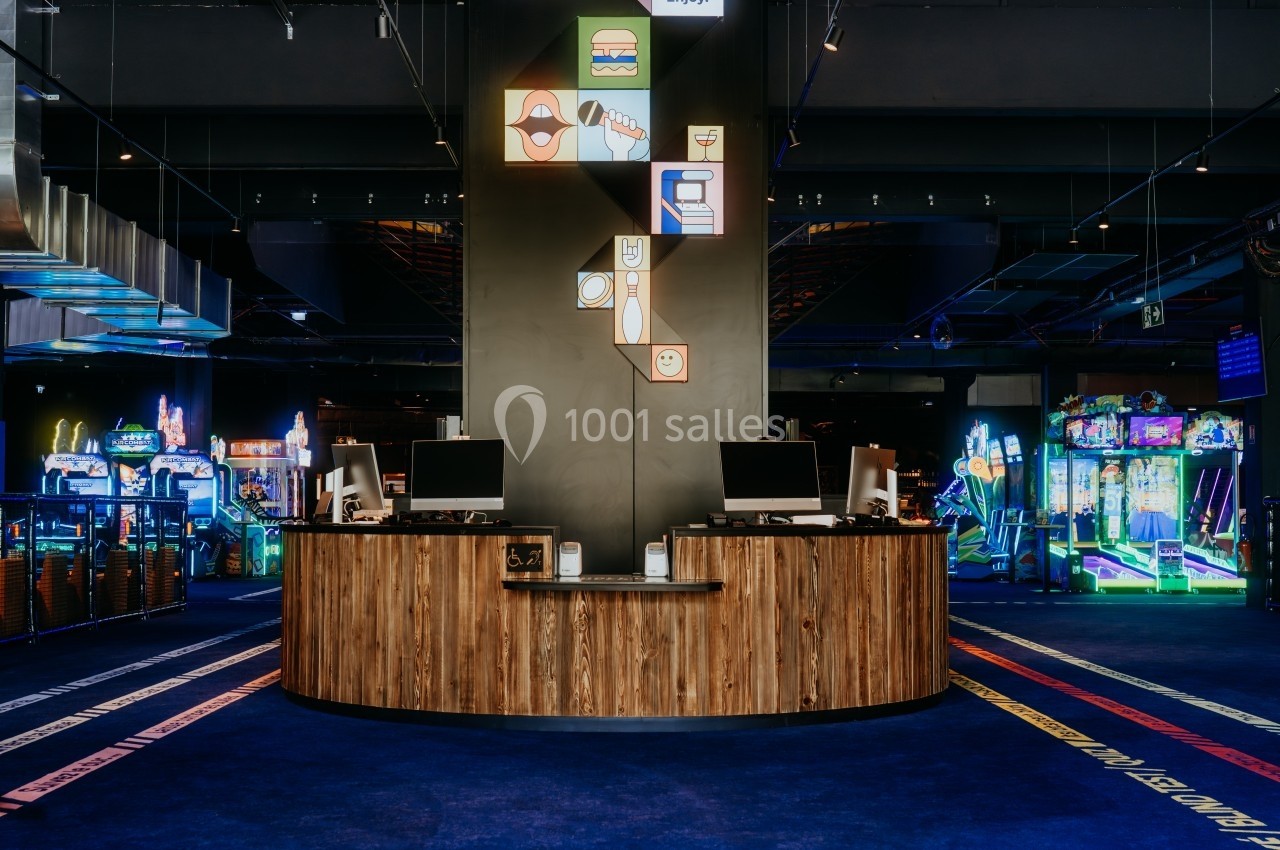 Comptoir en bois avec deux ordinateurs dans une salle d'arcade lumineuse, entourée de jeux et de machines.