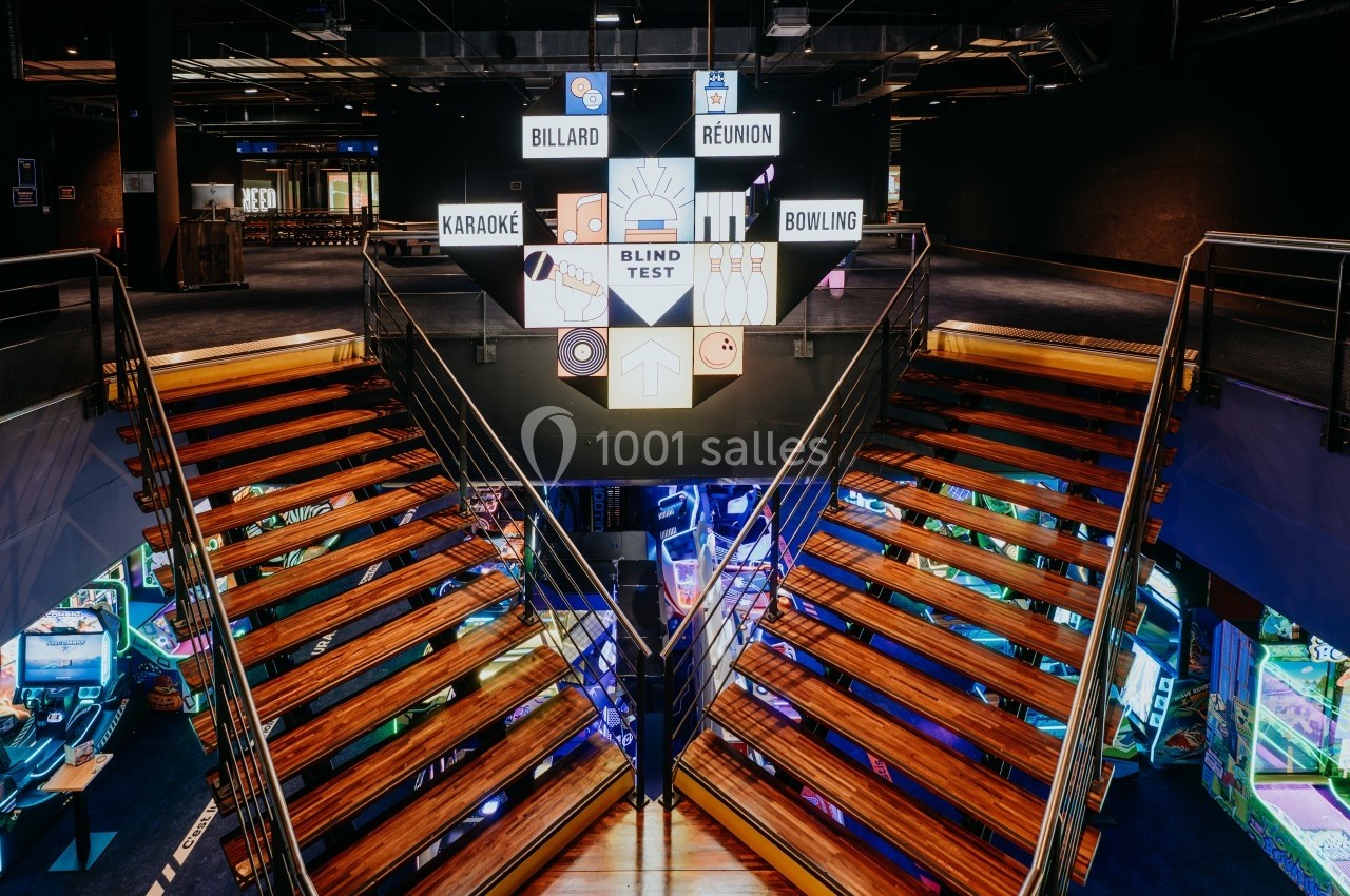 Escalier central en bois menant à des espaces de loisirs, avec des panneaux lumineux indiquant diverses activités.