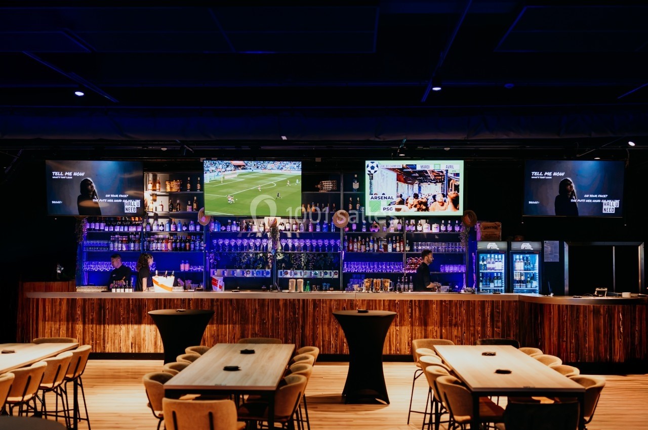 Bar moderne avec comptoir en bois, écrans diffusant des vidéos, étagères de bouteilles et tables en bois clair.
