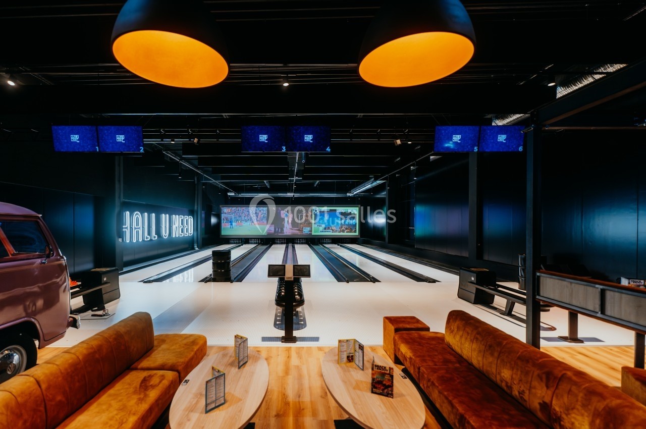 Salle de bowling moderne avec plusieurs pistes, canapés en velours, tables basses et écrans suspendus.