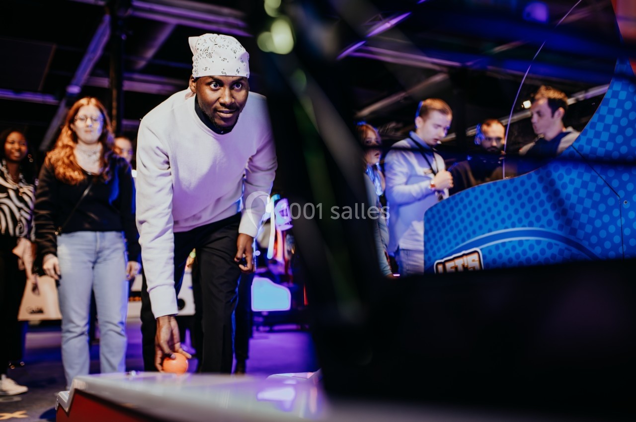 Un homme joue à un jeu d'arcade dans une salle animée, entouré de spectateurs.