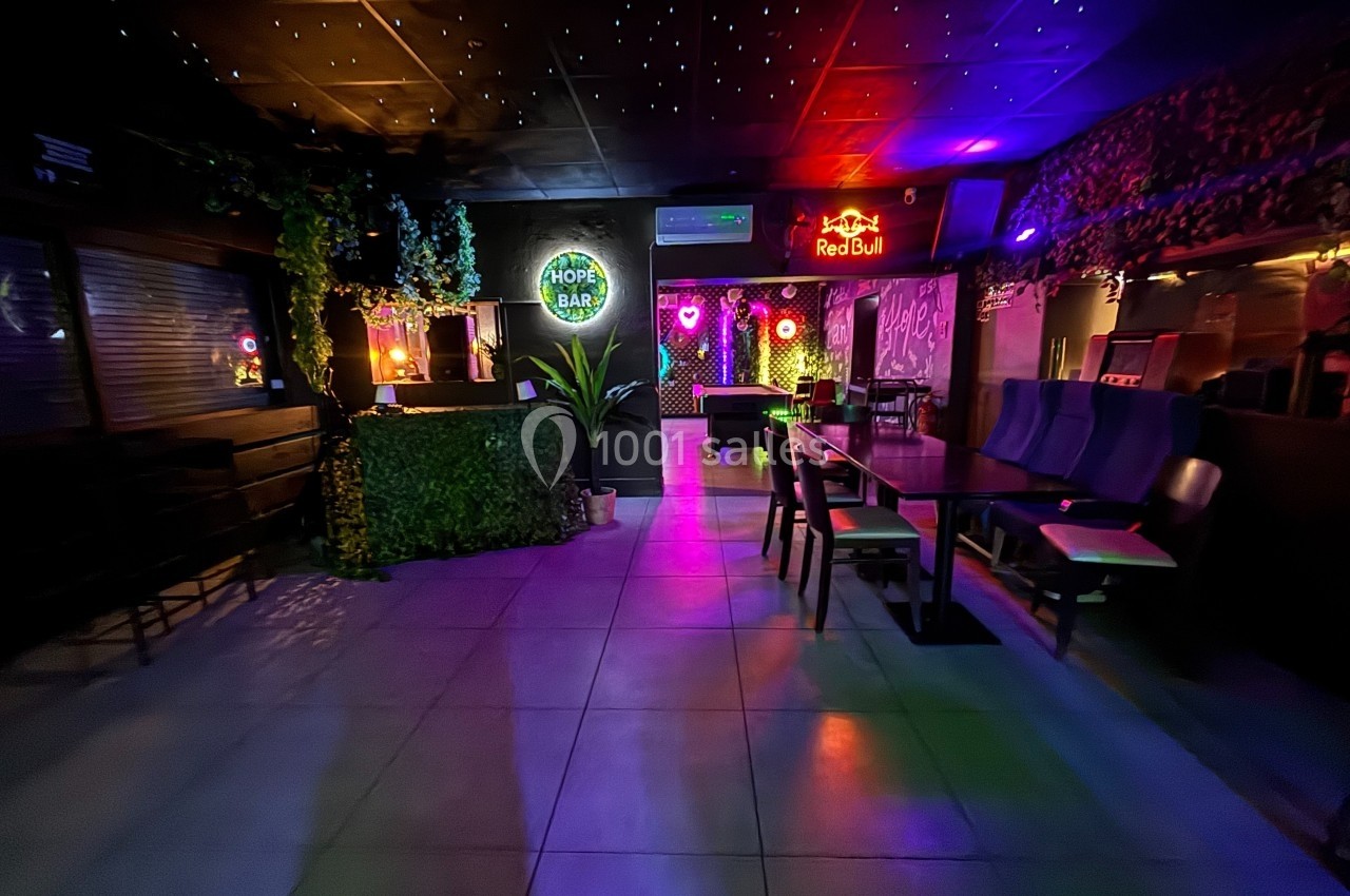 Salle de bar avec éclairage tamisé, néons colorés, plantes décoratives et mobilier moderne.