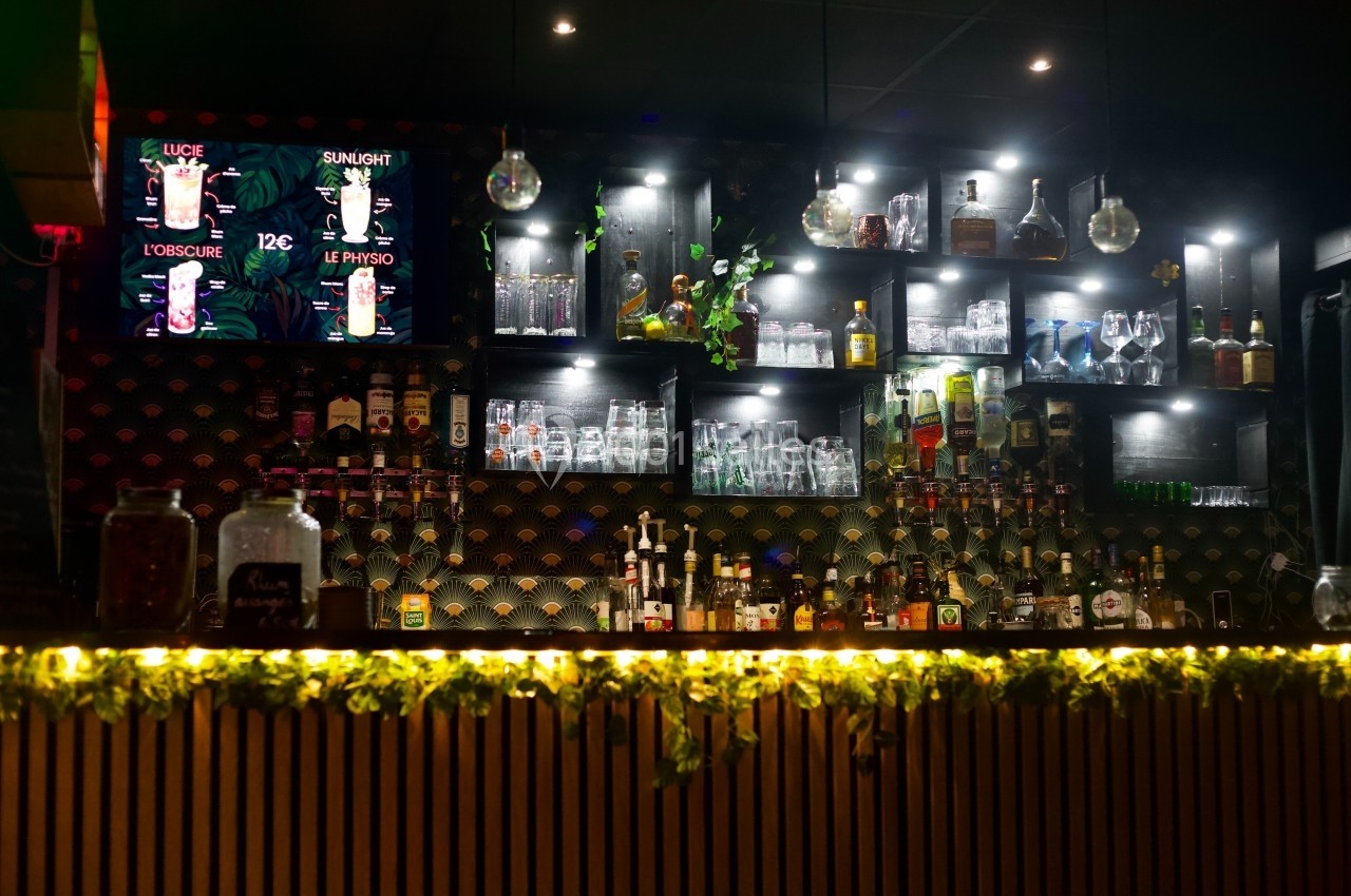 Bar avec étagères illuminées, bouteilles d'alcool, verres et écrans affichant des cocktails sur fond sombre.