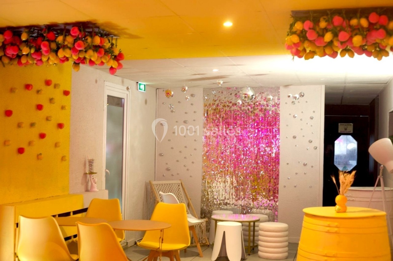 Salle lumineuse décorée de fleurs suspendues, mobilier jaune et blanc, et rideau scintillant rose en arrière-plan.
