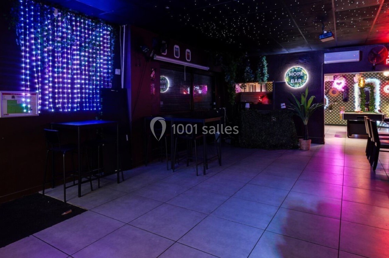 Salle de bar avec éclairage coloré, tables hautes, chaises noires et décorations lumineuses au mur.