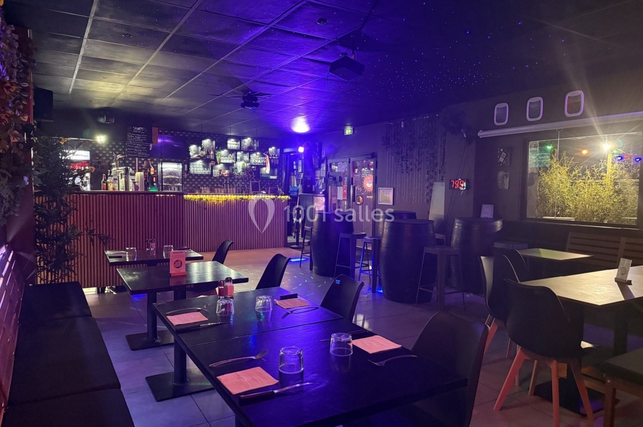 Salle de restaurant avec tables dressées, éclairage tamisé et bar illuminé en arrière-plan.