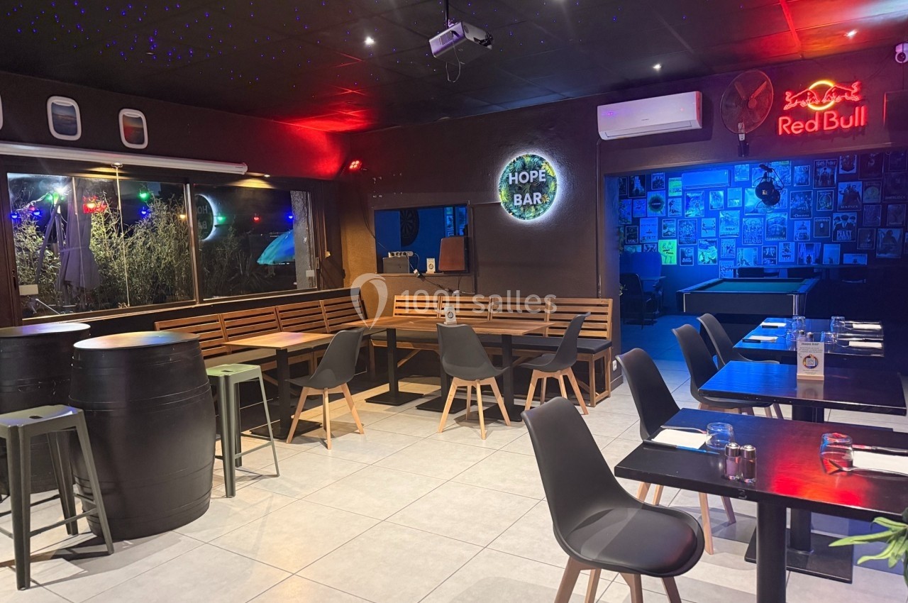 Salle de restaurant moderne avec tables, chaises, barils en guise de tables et enseignes lumineuses décoratives.