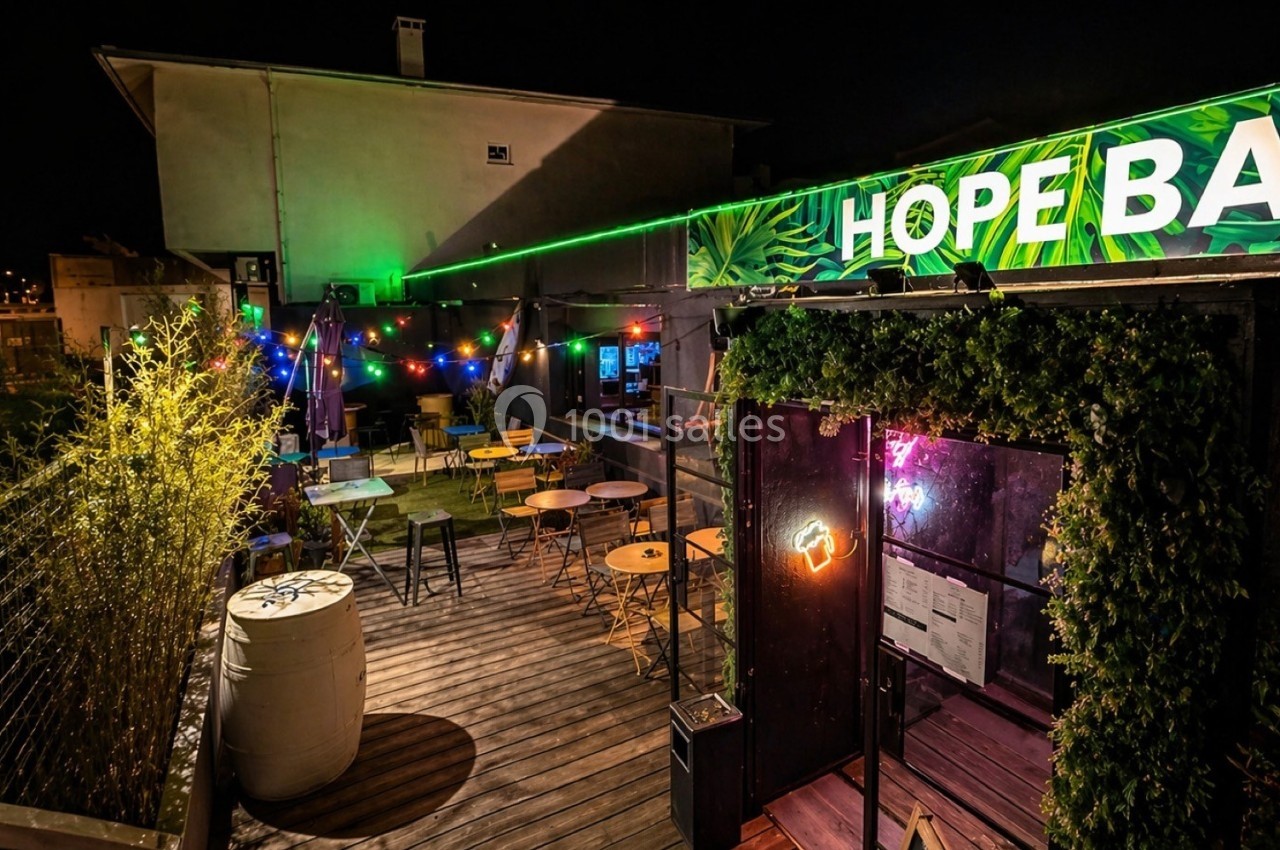 Terrasse extérieure d'un bar éclairée de guirlandes lumineuses colorées, avec tables et chaises disposées.