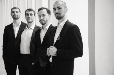 Miniature DJ Saint-Gilles (Bruxelles-Capitale) - 441 Quartet #6 Quatre musiciens en costume saluent sur une scène éclairée, entourés de bougies, dans un espace intérieur industriel.