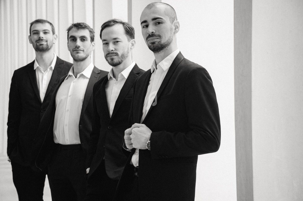 Quatre hommes en costume posent debout dans un espace lumineux avec des colonnes en arrière-plan.