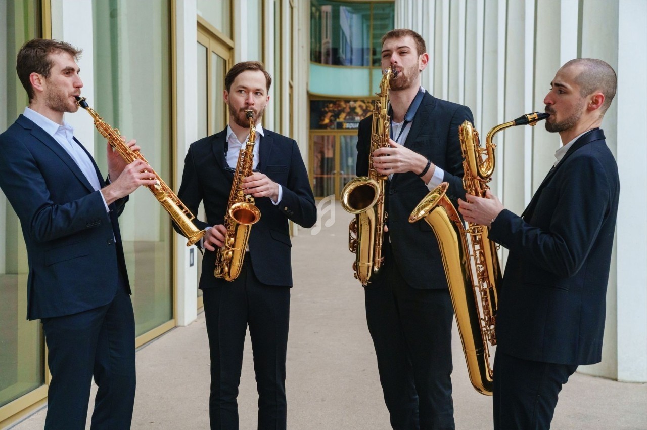 Quatre musiciens en costume jouent du saxophone devant un bâtiment moderne avec de grandes baies vitrées.