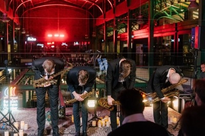 Miniature DJ Saint-Gilles (Bruxelles-Capitale) - 441 Quartet #10 Quatre musiciens en costume saluent sur une scène éclairée, entourés de bougies, dans un espace intérieur industriel.