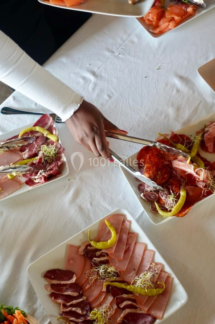 Une main sert des tranches de charcuterie variée sur une table dressée avec plusieurs assiettes de viandes froides.