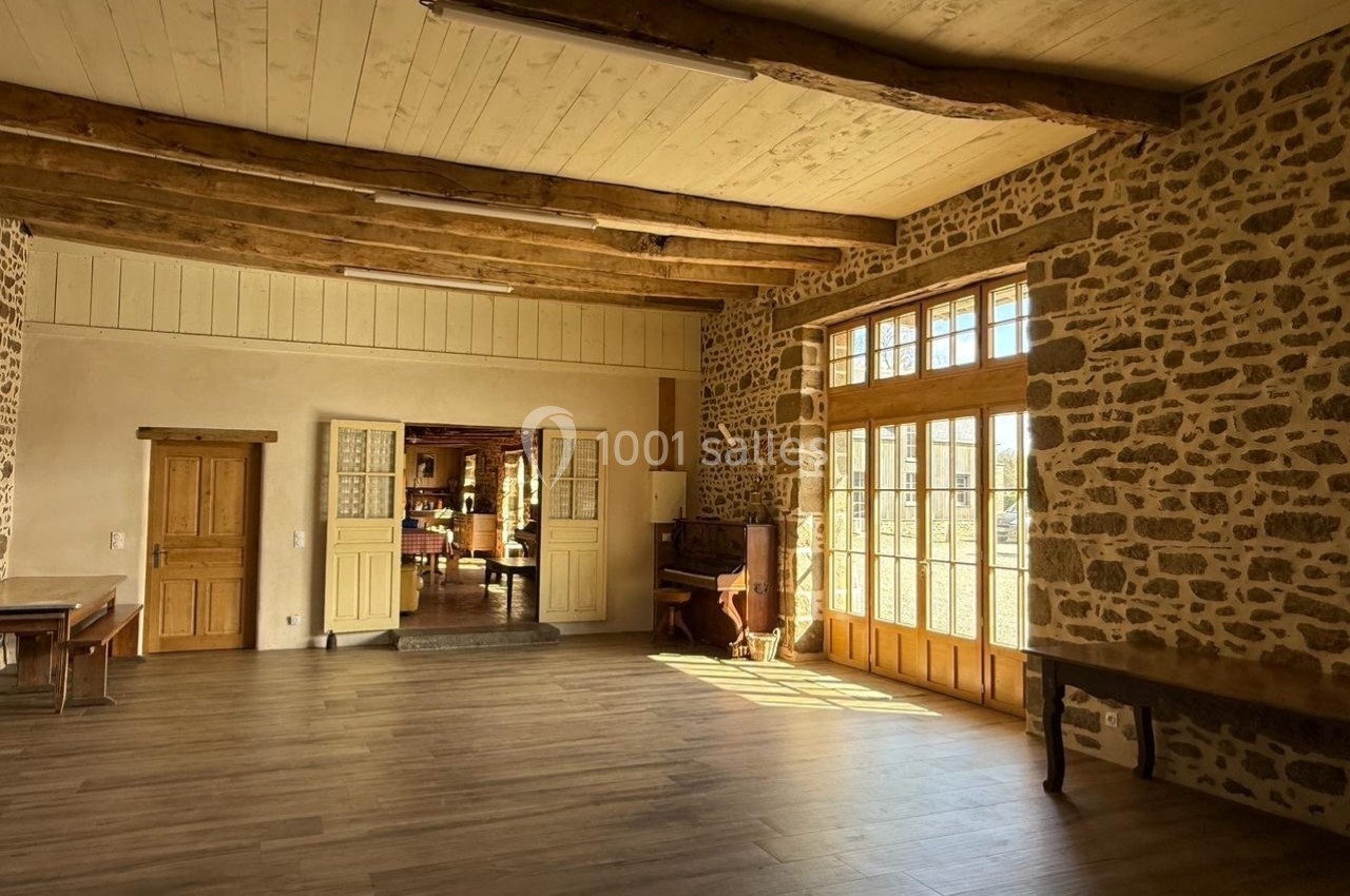 Salle lumineuse avec murs en pierre, poutres apparentes, parquet en bois et grandes portes vitrées donnant sur l'extérieur.