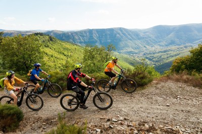 Quatre cyclistes en VTT sur un sentier de montagne entouré de végétation avec une vue sur une vallée boisée.