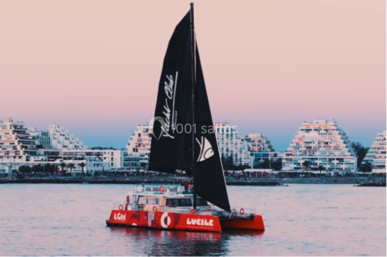 Catamaran rouge avec voiles noires naviguant près de bâtiments pyramidaux au bord de l'eau au coucher du soleil.