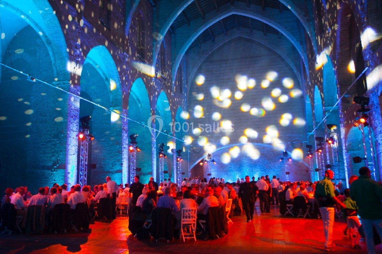 Salle voûtée illuminée par des projections colorées, avec des tables dressées et des convives assis ou debout.