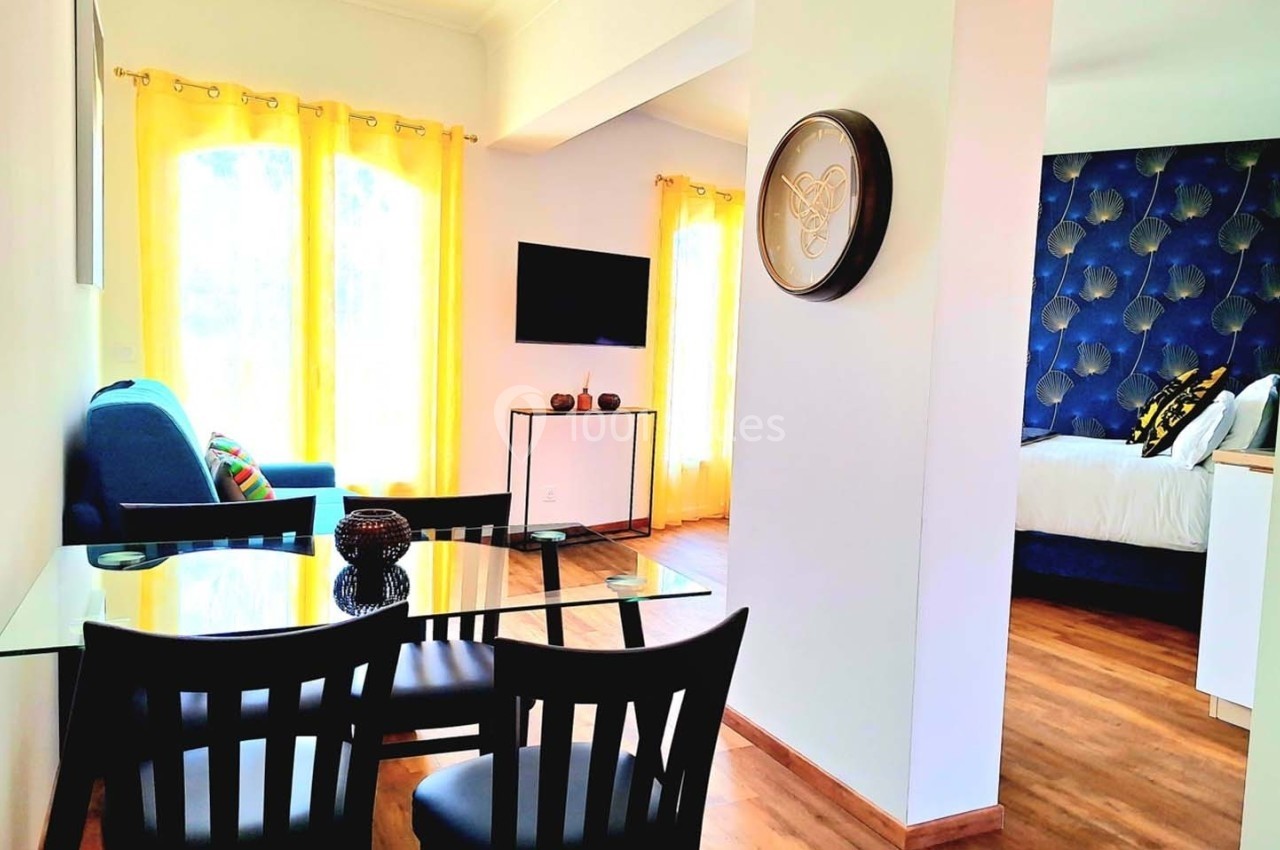 Salle à manger avec table en verre et chaises noires, salon avec canapé bleu et chambre avec lit à tête capitonnée bleue.
