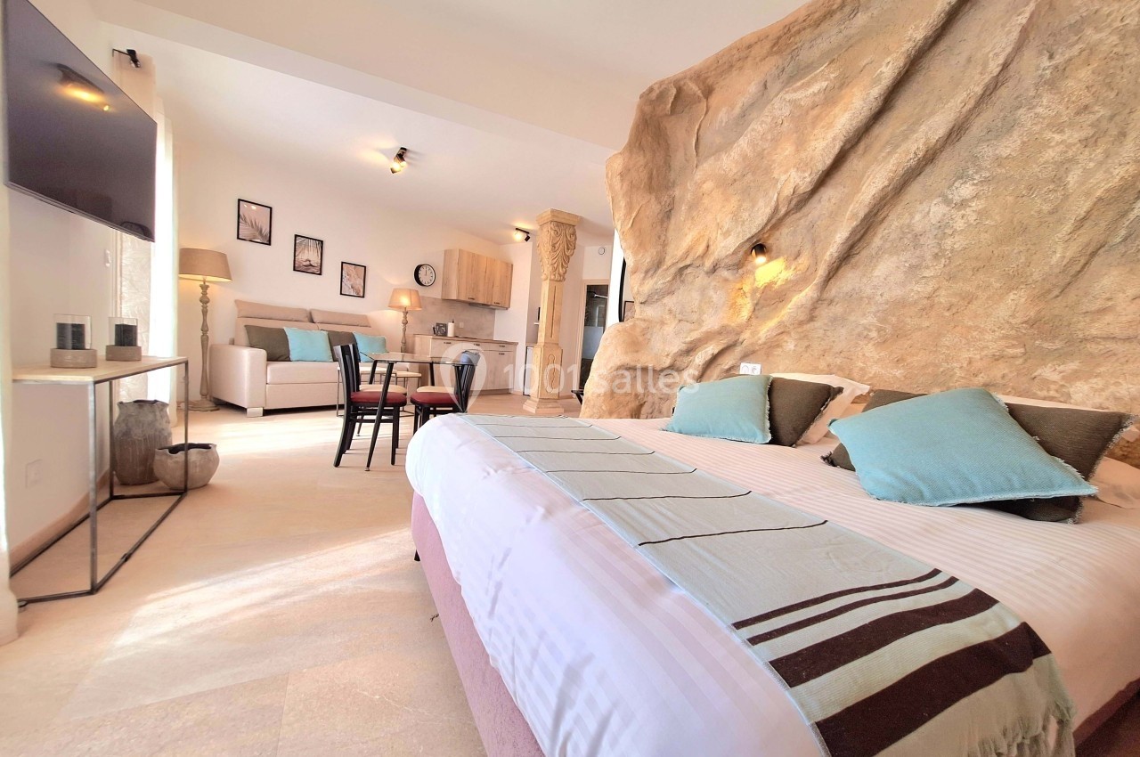 Chambre lumineuse avec lit double, mur en pierre naturelle, coin salon et cuisine ouverte en arrière-plan.